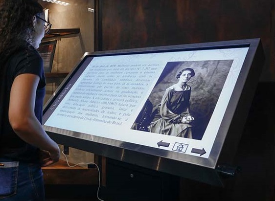 Mulher acessando a exposição virtual em tela interativa