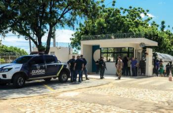 Pessoas estão reunidas em frente a uma guarita, com uma viatura da Guarda Municipal estacionada ao lado, em um ambiente arborizado e ensolarado.