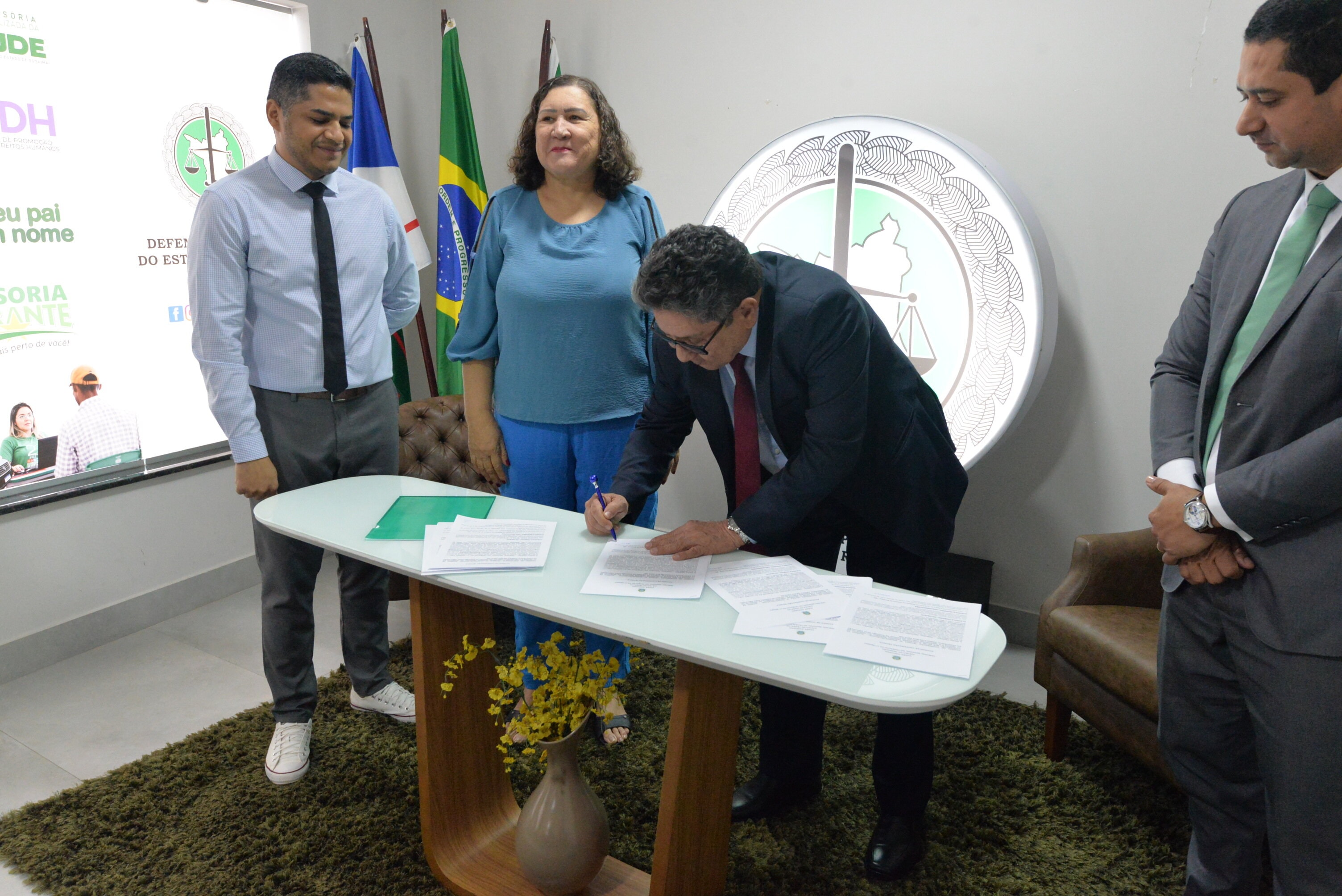Fotografia com 4 pessoas presentes, sendo 3 homens e 1 mulher. Um deles está assinando um papel sobre a mesa.