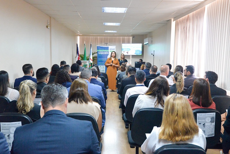 A imagem mostra um auditório cheio de pessoas assistindo a uma apresentação. Ao fundo, há duas mulheres em pé, uma delas falando ao público. Banners, bandeiras e uma tela com informações compõem o cenário. A atmosfera é formal, sugerindo um evento corporativo ou institucional.