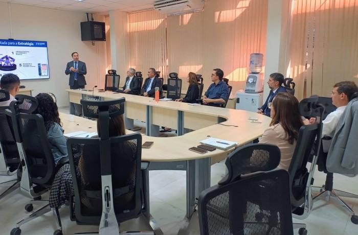 Pessoas participam de uma reunião em sala com mesa em U; um homem em pé apresenta slides em uma tela.