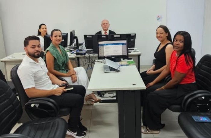 Fotografia com a presença de 6 pessoas, sendo 4 mulheres e 2 homens, 4 estão sentados e 2 no fundo da sala em frente a computadores. 