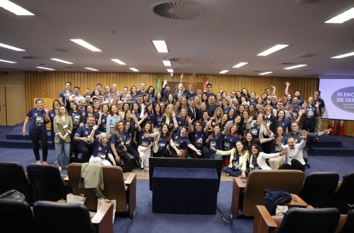 A imagem mostra os participantes do encontro posando para foto em auditório fechado.