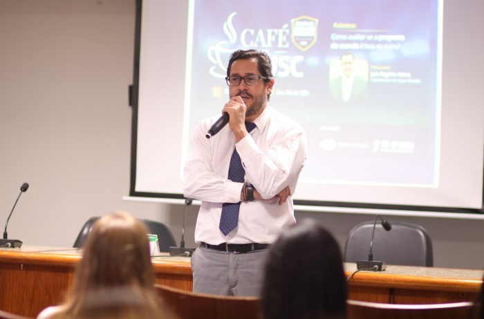 A imagem mostra o magistrado palestrante com projeção da apresentação ao fundo.