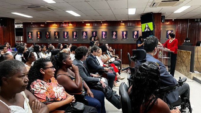 A imagem mostra uma mulher falando em um púlpito em uma sala cheia de pessoas sentadas, ouvindo atentamente. Ao fundo, há retratos na parede e uma tela com intérprete de Libras.