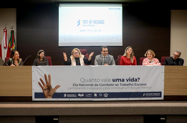 Fotografia feita em um ambiente fechado, com 7 pessoas centralizadas e sentadas no palco do auditório do TRT-8 (PR) e o cartaz logo abaixo escrito ''Quanto vale uma vida?'' em destaque.