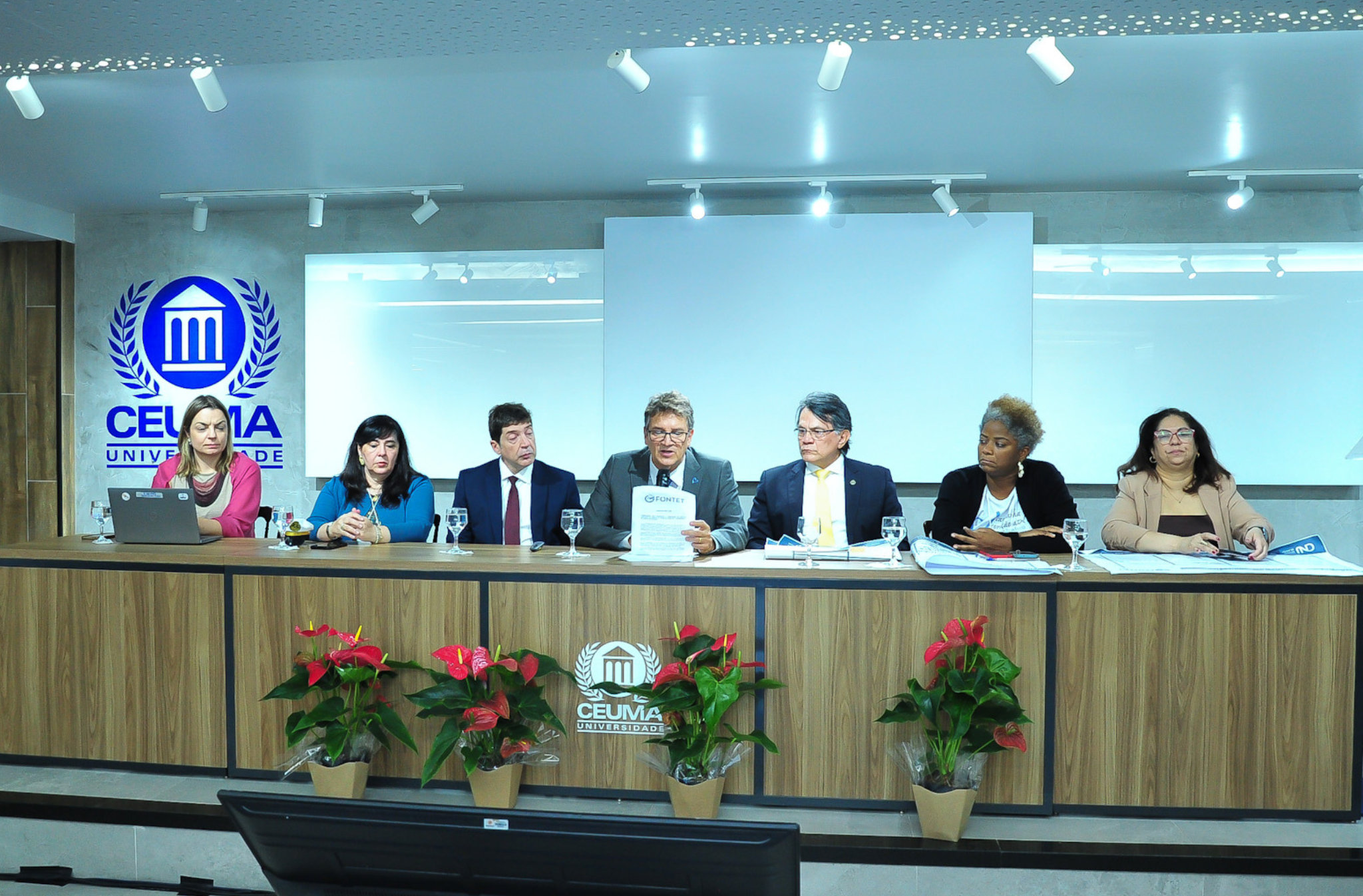 A imagem mostra uma sala com sete pessoas sentadas a uma mesa. No centro da mesa, um homem segura um microfone e lê um papel.