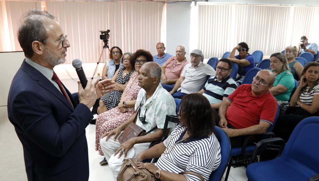 Fotografia de um homem com o microfone na mão falando com algumas pessoas que estão sentadas em cadeiras azuis. 