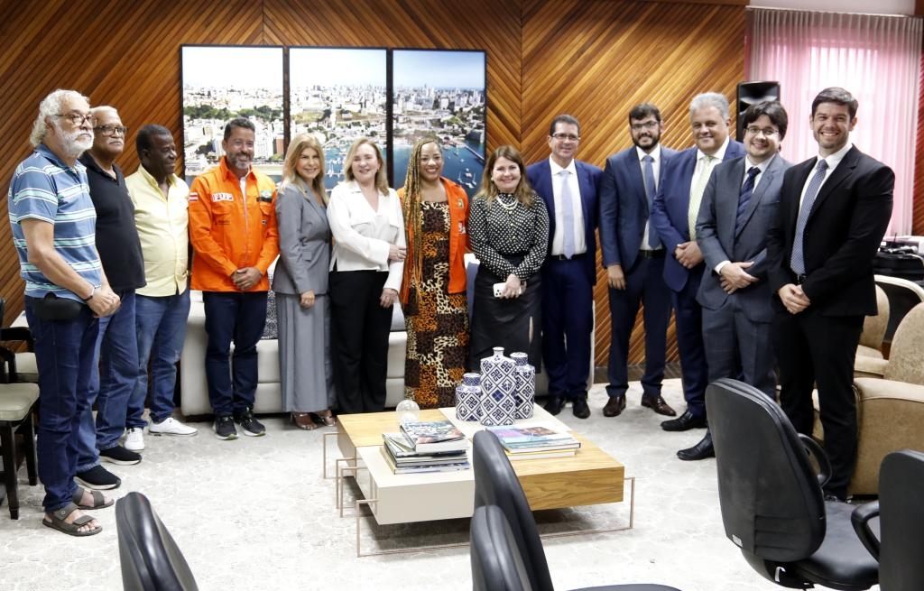 A imagem mostra um grupo de pessoas posando para foto em uma sala de reuniões. Elas estão em pé, sorrindo, com uma mesa de centro à frente e quadros com vista urbana ao fundo.
