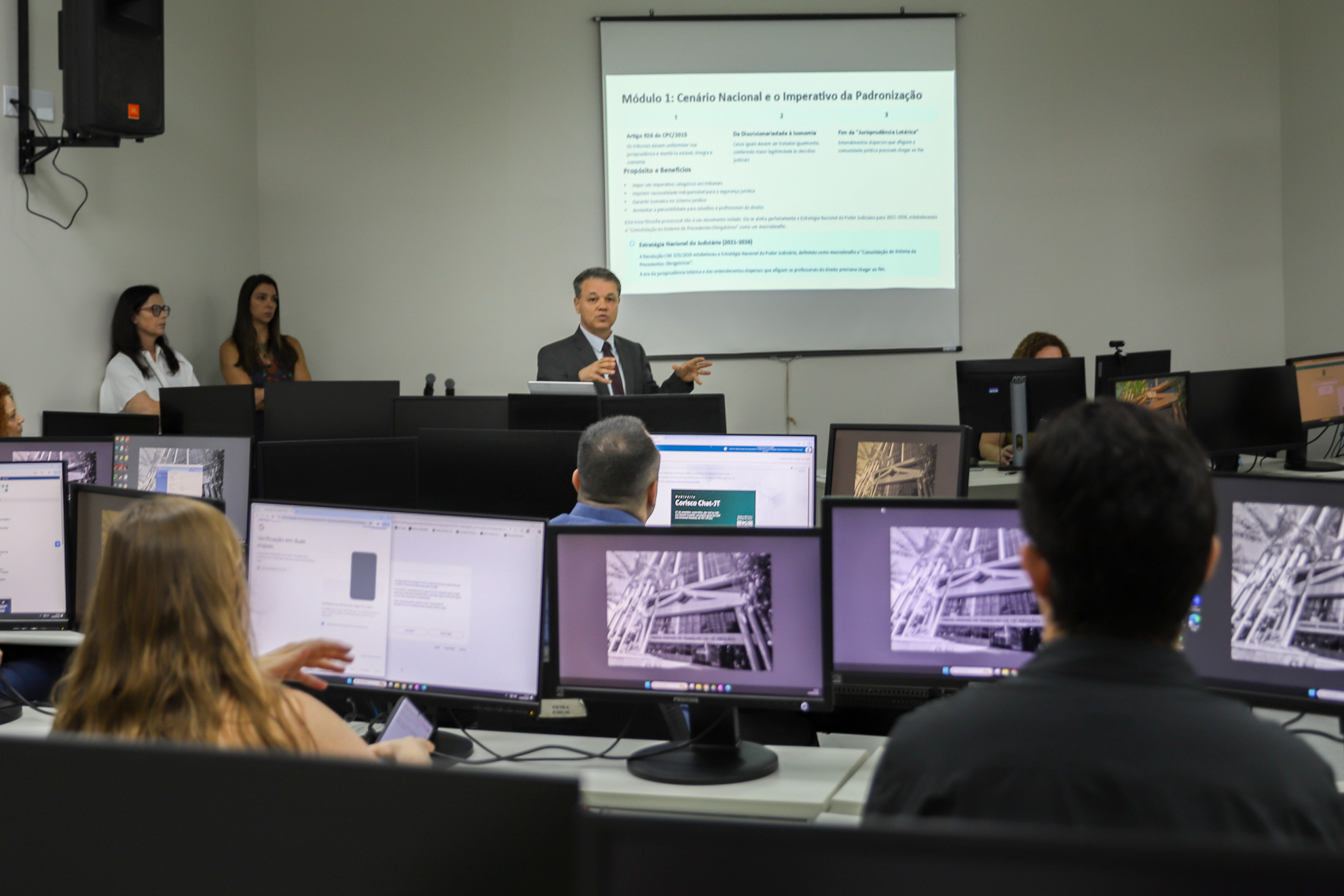 Imagem mostra uma sala de treinamento, com pessoas em frente a computadores enquanto um homem, em pé, faz uma apresentação usando projetor.