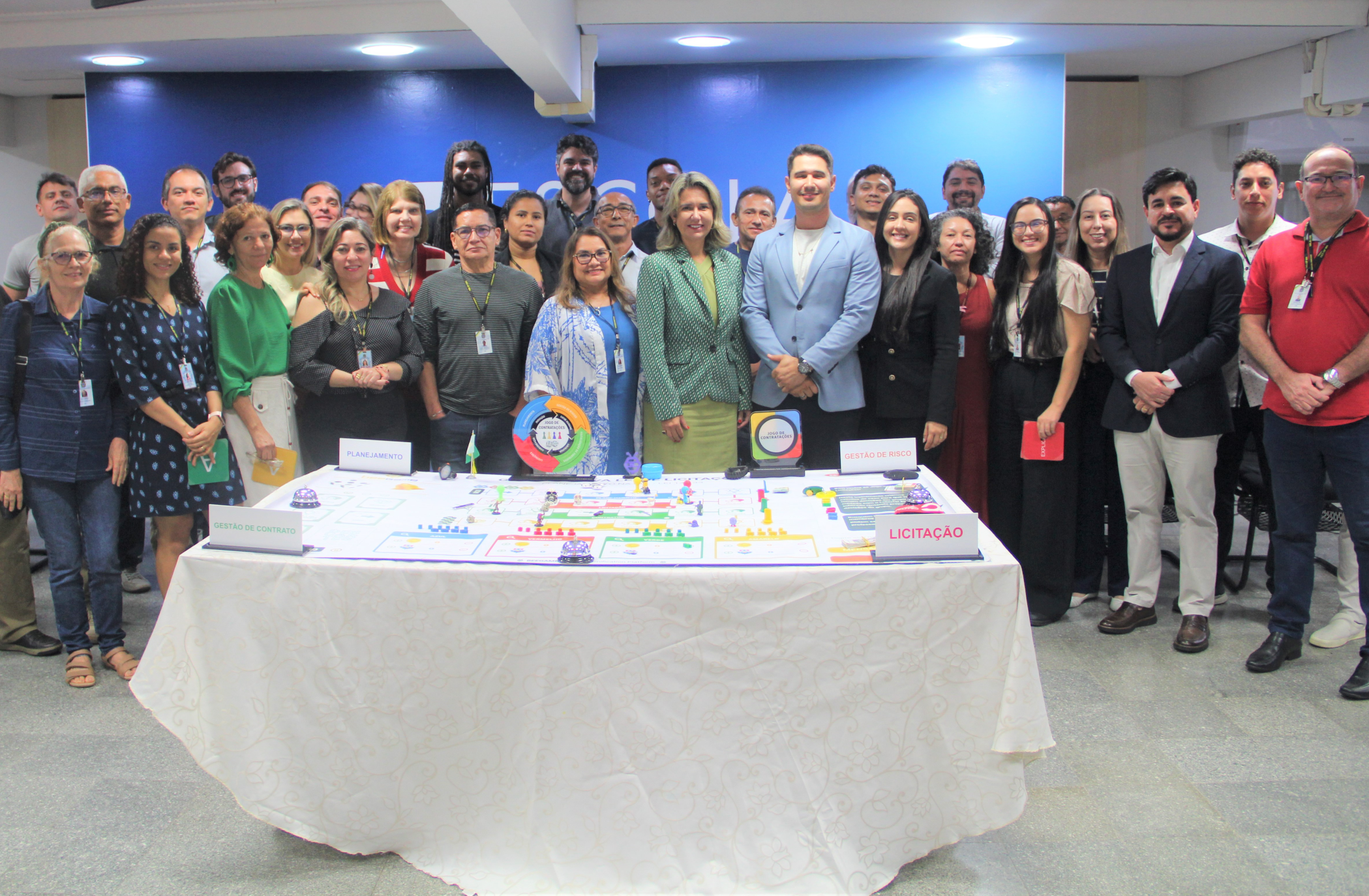 Foto de várias pessoas de pé e sorrindo. À frente delas, mesa com toalha branca com peças de um jogo distribuídas em cima.