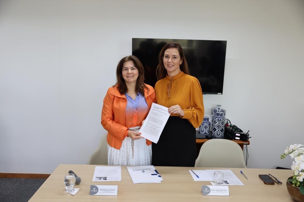 Fotografia com a presença de duas mulheres, ambas segurando um papel e há alguns papeis sobre a mesa.