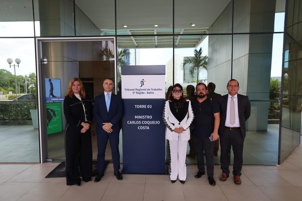Fotografia tirada com 5 pessoas, 3 do lado direito, 2 homens e 1 mulher; 2 do lado esquerdo, 1 mulher e 1 homem. Ao meio, uma placa do TRT 5 (BA) Torre 02 Ministro Carlos Coqueijo Costa.