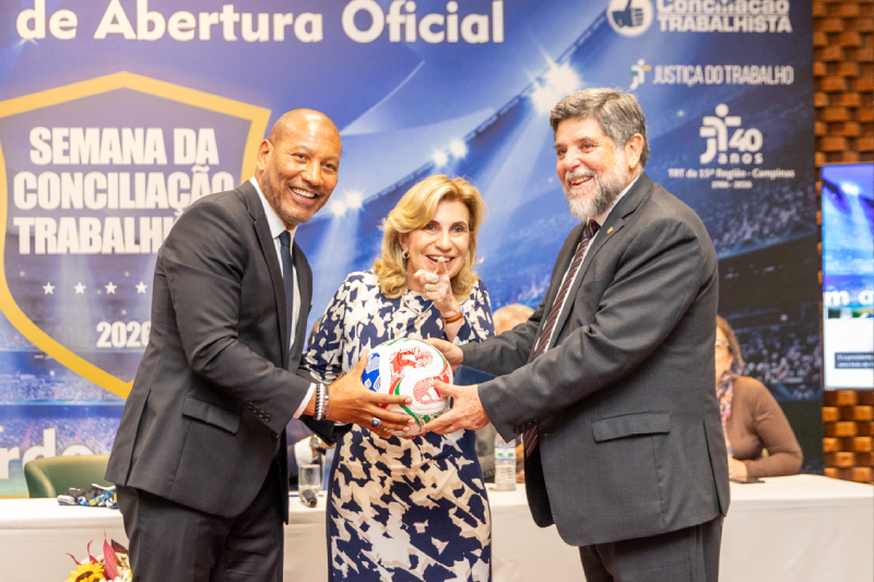 Tetracampeão Mauro Silva, desembargadora Ana Paula Lockmann e ministro Caputo segurando uma bola da Copa 2026 com o banner da semana ao fundo.