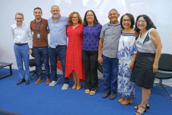 Grupo de oito pessoas posando sorridentes lado a lado em uma sala com carpete azul e cadeiras ao fundo.