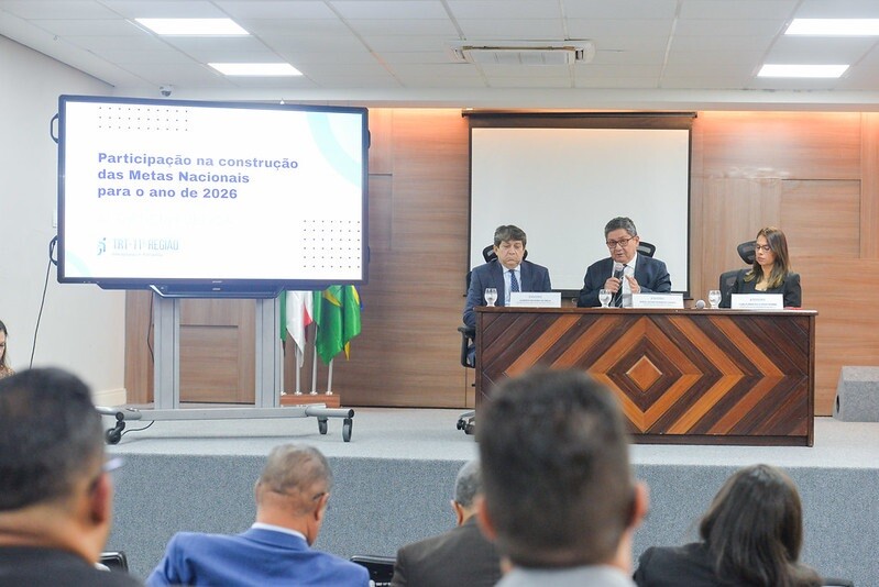 Fotografia com 3 pessoas sentadas palestrando e, ao lado, um banner com a legenda ''Participação na construção das Metas Nacionais para o ano de 2026''.