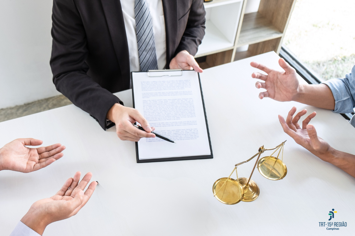 Uma mão segura uma prancheta com um documento e uma caneta, enquanto as outras gesticulam. Sobre a mesa há uma balança dourada e no canto inferior direito aparece o logotipo do TRT-15ª Região – Campinas.