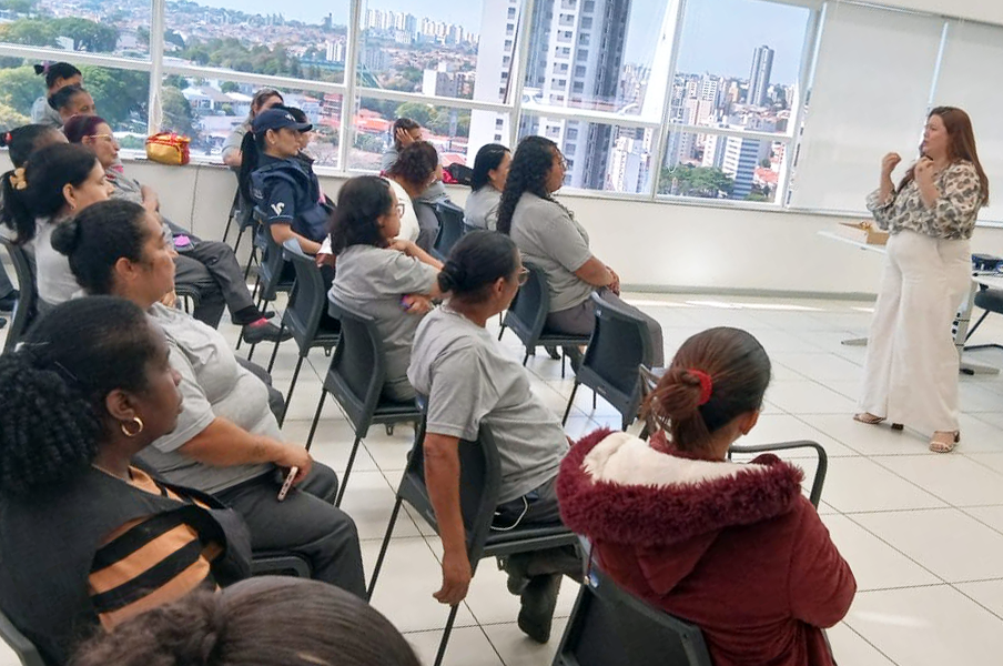 Fotografia com mulheres sentadas em diversas cadeiras e uma mulher em pé falando algo.