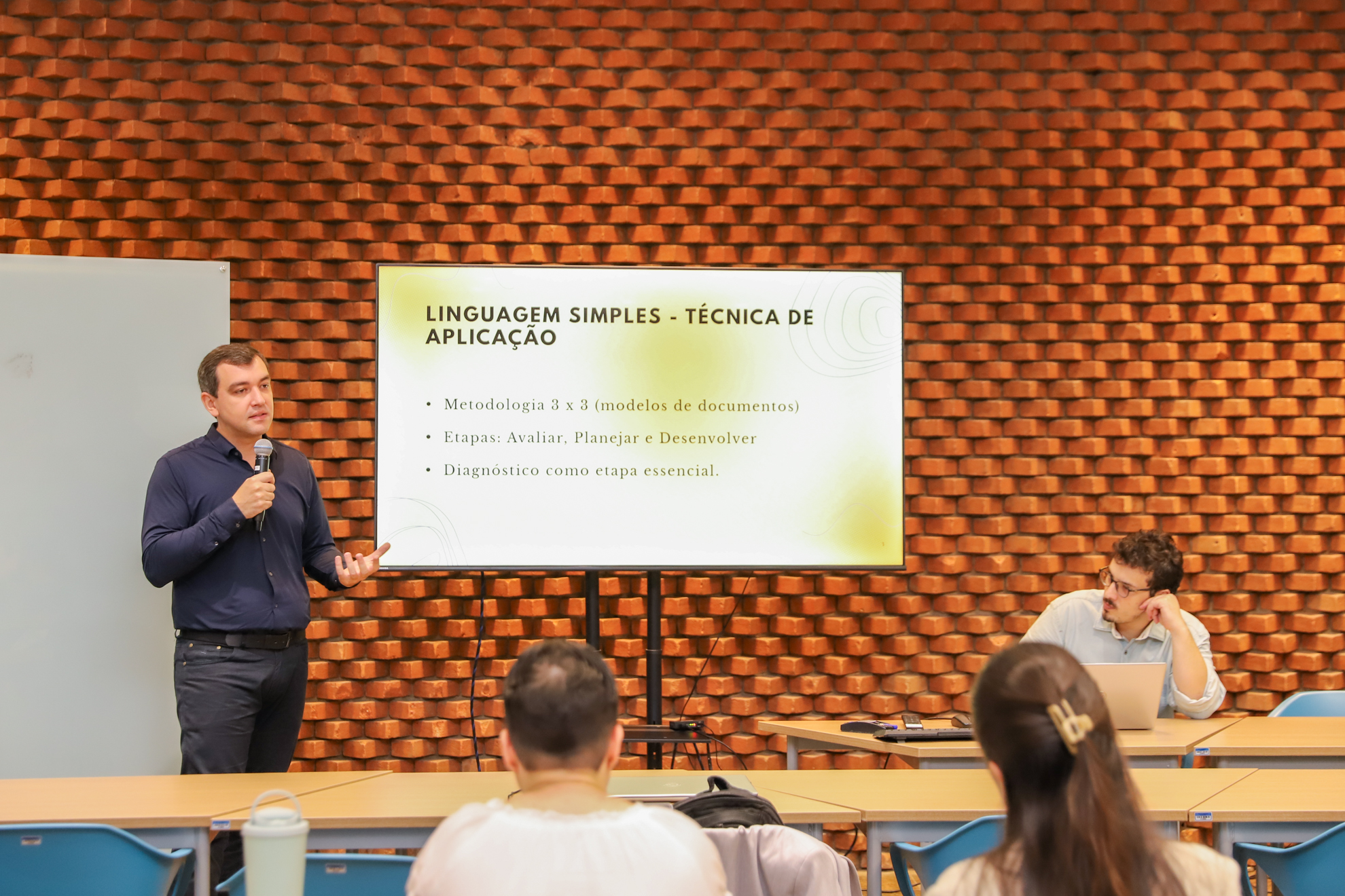 Fotografia com um homem segurando microfone, ao lado de um banner com o texto: Linguagem simples - técnica de aplicação Metodologia 3 x 3 (modelos de documentos) Etapas: Avaliar, Planejar e Desenvolver Diagnóstico como etapa essencial.