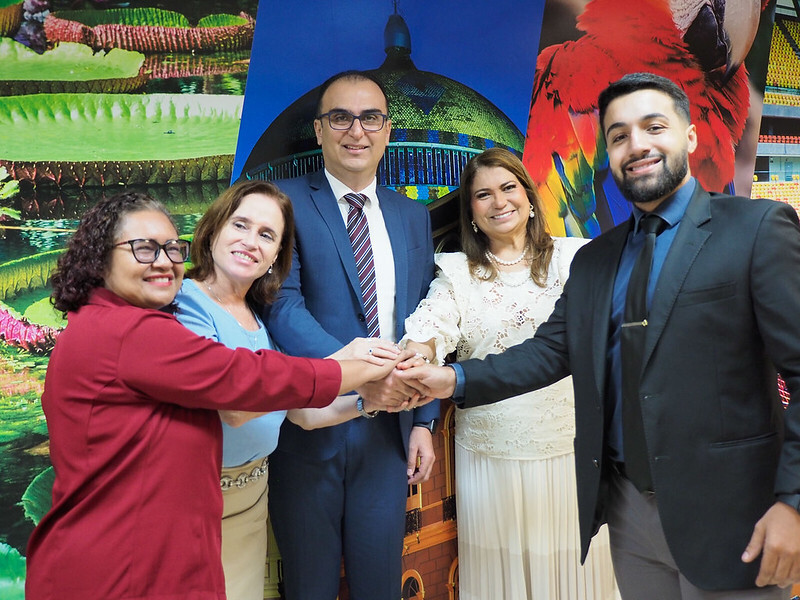 A imagem mostra cinco pessoas sorrindo e unindo as mãos ao centro, em gesto de união. Ao fundo, há painéis coloridos com imagens de paisagens, um teatro e uma arara.