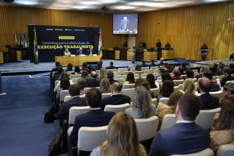 Foto do plenário com a cadeiras ocupadas com os participantes, além da mesa com os três ministros durante a abertura. Ao fundo dos ministros, um backdrop com a identidade visual do evento.