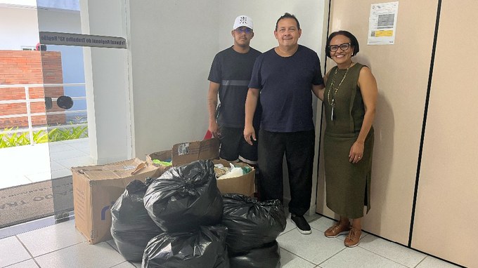 Fotografia com a presença de 3 pessoas, 2 homens e 1 mulher; a frente deles algumas caixas e sacolas pretas. 