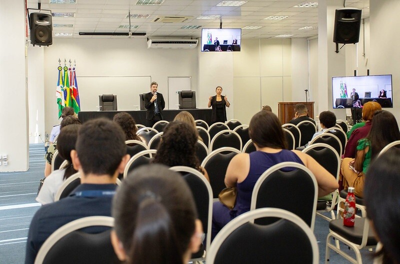 Palestra acontecendo em um auditório com cadeiras ocupadas. Um homem fala ao microfone no palco e uma mulher faz interpretação em libras. Há bandeiras ao fundo e telões exibindo a apresentação.