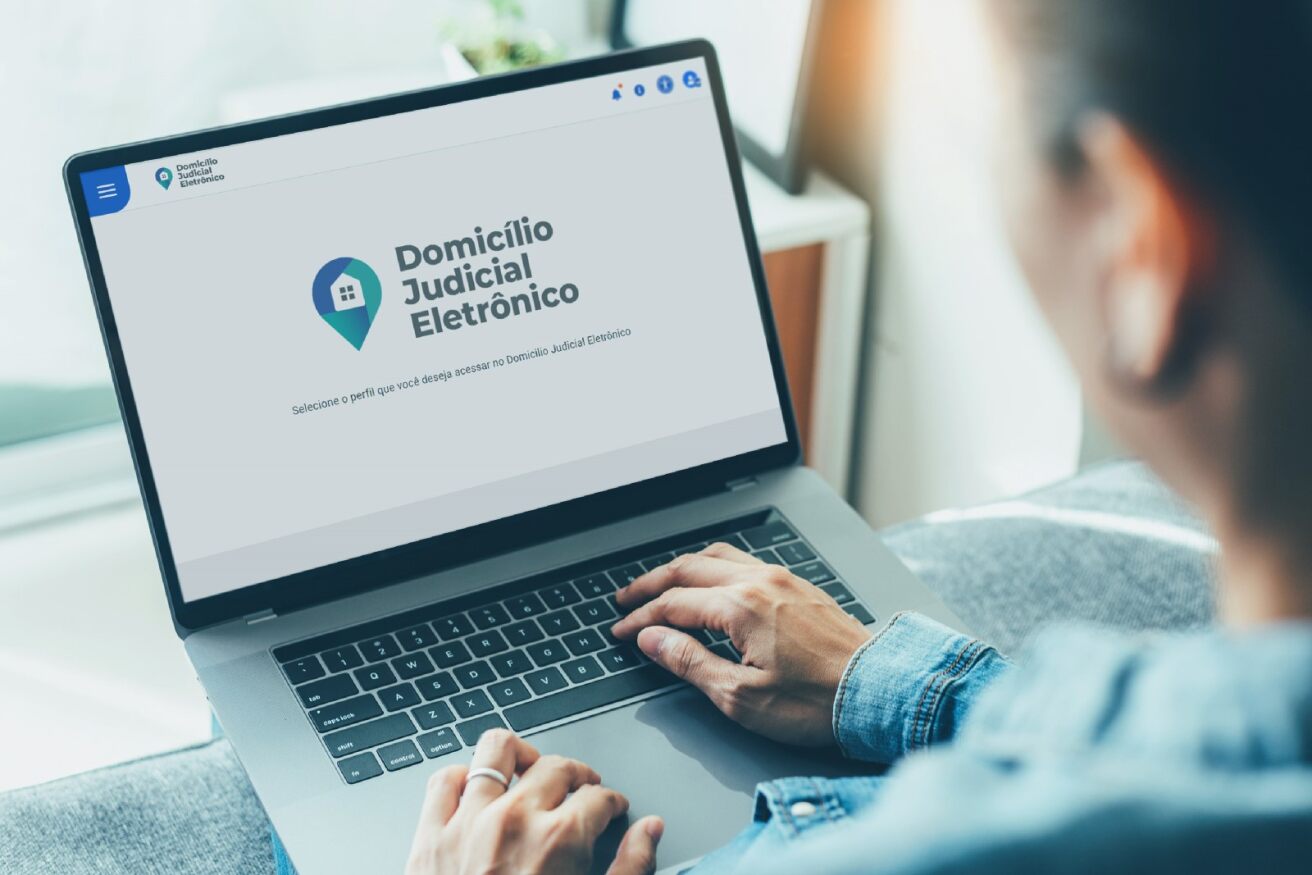 Foto de pessoa com notebook no colo e mãos sobre o teclado. Na tela, a logo do Domicilio Judicial Eletrônico