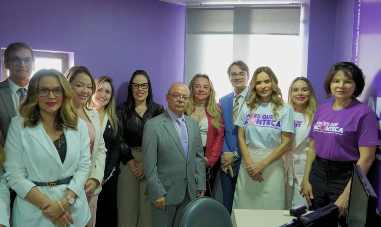 A imagem mostra um grupo de pessoas posando para foto em uma sala com paredes roxas. Algumas usam camisetas com a frase 