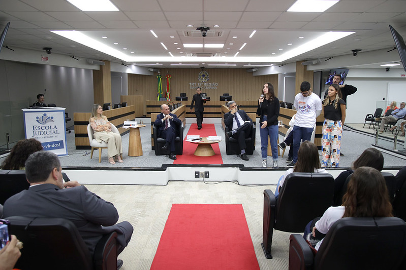 Uma adolescente falando no microfone. Ministro Lelio sentado em cadeira na frente da plateia observando a jovem falar.