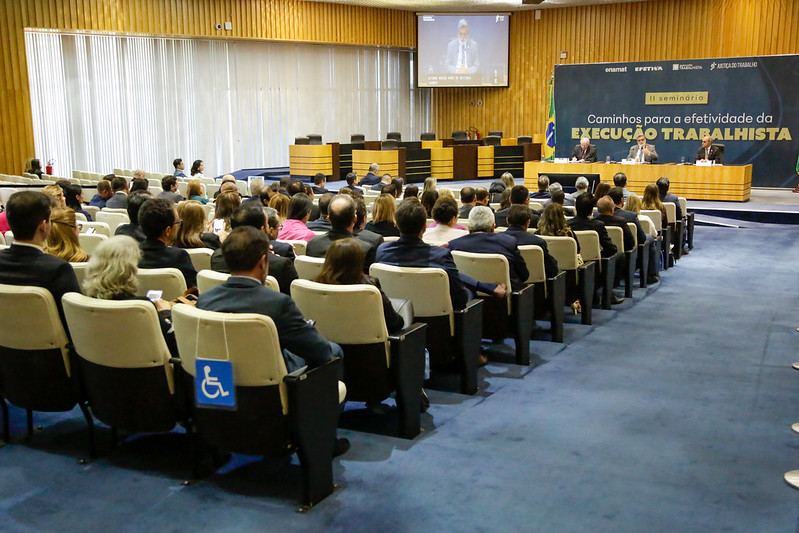 Foto do plenário do TST com pessoas sentadas nas cadeiras e, no palco, uma mesa de abertura com os ministros Lelio Bentes, Mauricio Godinho e Cláudio Brandão 