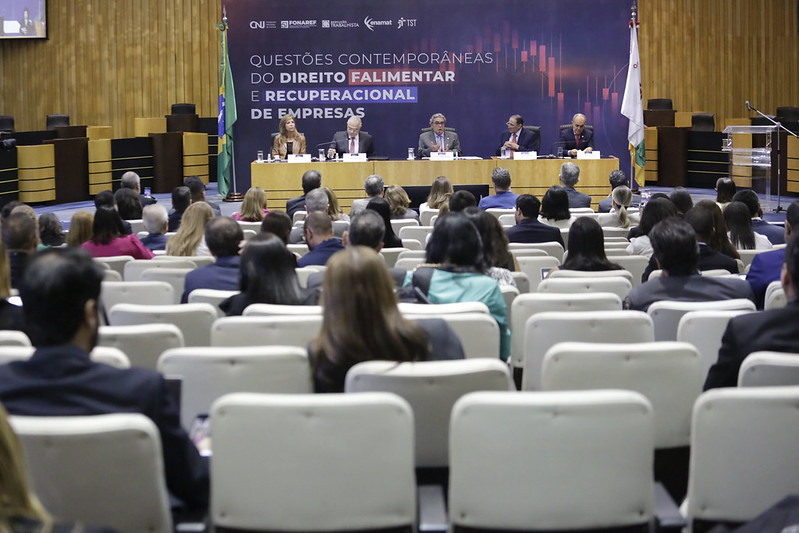 Foto do plenário com pessoas sentadas olhando a mesa de abertura do evento, que tem um backdrop ilustrativo do evento. Na mesa tem cinco pessoas.