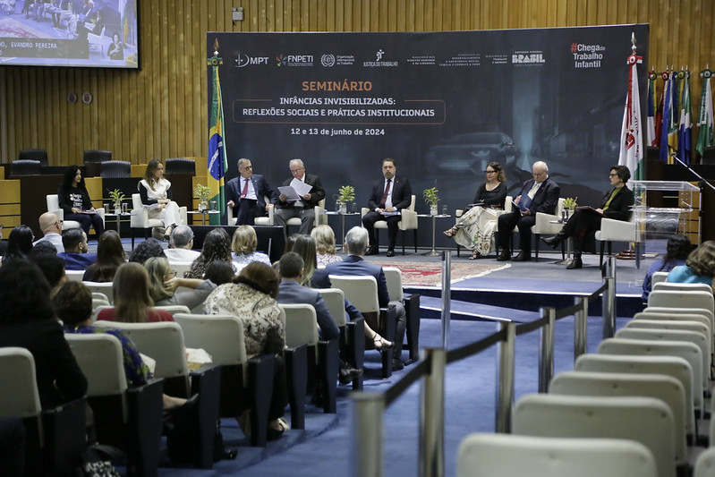 Foto do plenário do TST com pessoas sentadas e um backdrop do seminário ao fundo. na frente do backdrop estão as pessoas sentadas em cadeiras para a abertura do evento.