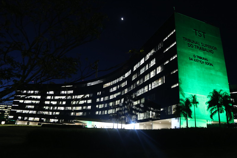 Foto da fachada do TST e do CSJT iluminada na cor verde. Foto a noite, com céu estrelado e o prédio com um paredão de concreto esverdeado.