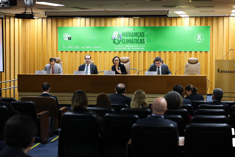 Foto do auditório com quatro pessoas na mesa. Ao fundo, um banner do evento predominantemente na cor verde.