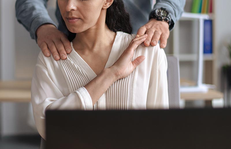 Mulher sentada recusa toque nos ombros feito por um homem atrás dela. Ela parece desconfortável. Cena sugere assédio no trabalho.