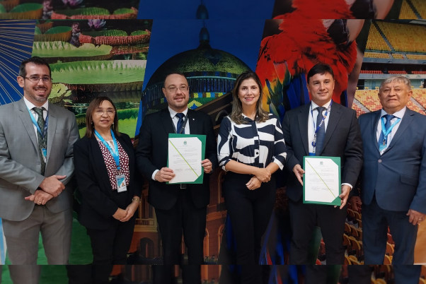 Grupo de quatro homens, dos quais dois seguram certificados, e duas mulheres com roupas formais, em pé. Ao fundo há um painel colorido com imagens amazônicas ao fundo.