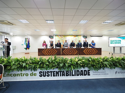 Fotografia com o banner ''Encontro de Sustentabilidade'' e uma mesa diretiva ao fundo com algumas pessoas em pé. 