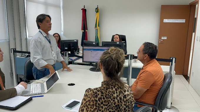A imagem mostra 5 pessoas em um ambiente de escritório, com um homem de camisa branca em pé gesticulando enquanto fala com outras 4 pessoas sentadas ao redor de uma mesa. Há computadores e documentos na mesa, e bandeiras em um mastro ao fundo.