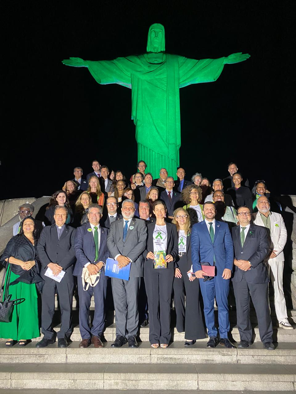 Diversas pessoas posadas para foto na escadaria aos pés do cristo redentor, que está na cor verde.