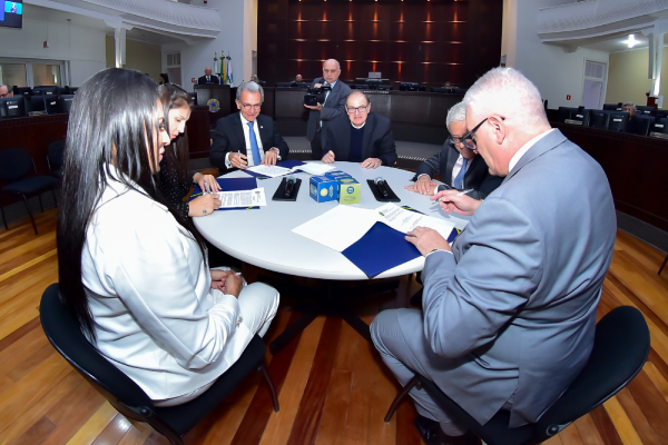 Mesa redonda com pessoas assinando papeis do acordo.