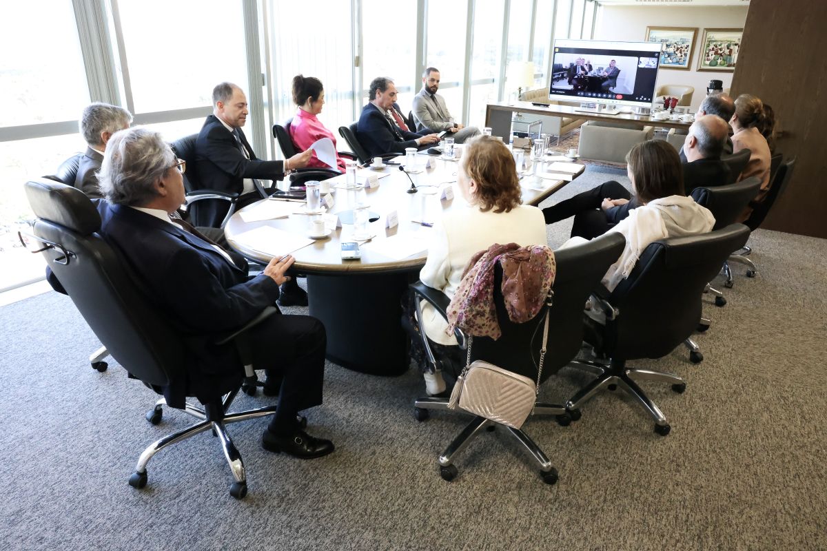 Fotografia de uma sala de reunião com 11 pessoas sentadas e um computador com a tela aberta em uma chamada de vídeo.