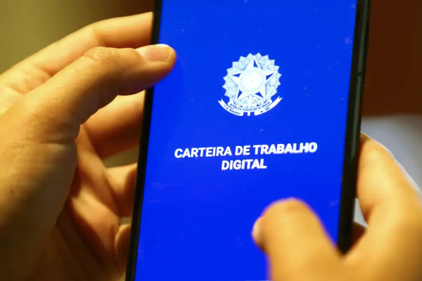 Foto de mãos segurando um celular. Na tela em fundo azul está escrito Carteira de Trabalho Digital em branco e o brasão da republica na mesma cor.
