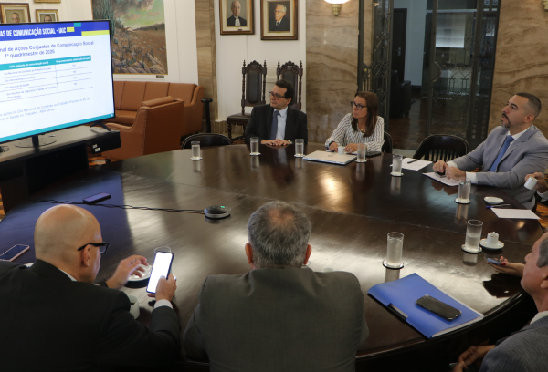 A imagem mostra um grupo de pessoas em uma reunião ao redor de uma mesa redonda, observando uma apresentação em uma tela.