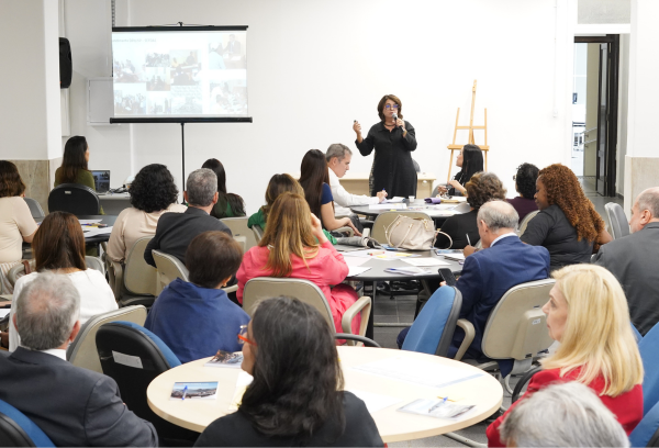 Palestrante fala ao público em uma sala com várias mesas e pessoas sentadas, enquanto uma apresentação é exibida em um telão ao fundo.