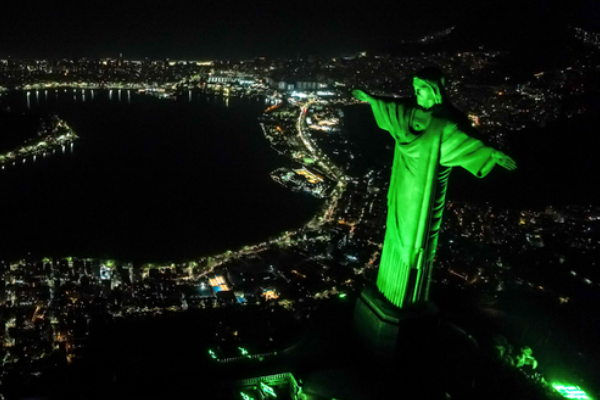 Foto aérea do Cristo Redentor iluminado de verde. Ao fundo, a cidade do Rio de Janeiro a noite.