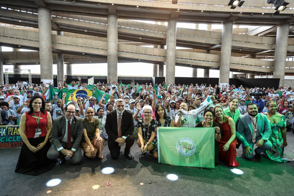 Várias pessoas agachadas, incluindo as autoridades e a categoria de catadores. Ao fundo uma parte da estrutura do estádio.