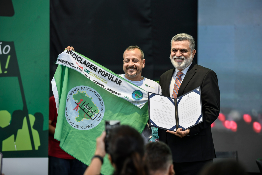Ministro posando ao lado de um representante do catadores. O ministro seguro o ato que instituiu o grupo de trabalho e o representante segura uma bandeira do movimento na cor verde. 