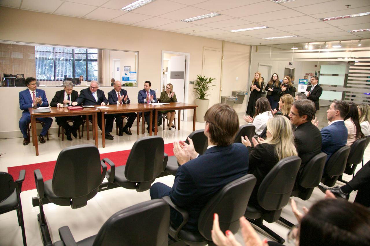 A imagem mostra uma cerimônia formal de assinatura de acordo, com autoridades sentadas à mesa e plateia aplaudindo em um ambiente corporativo.