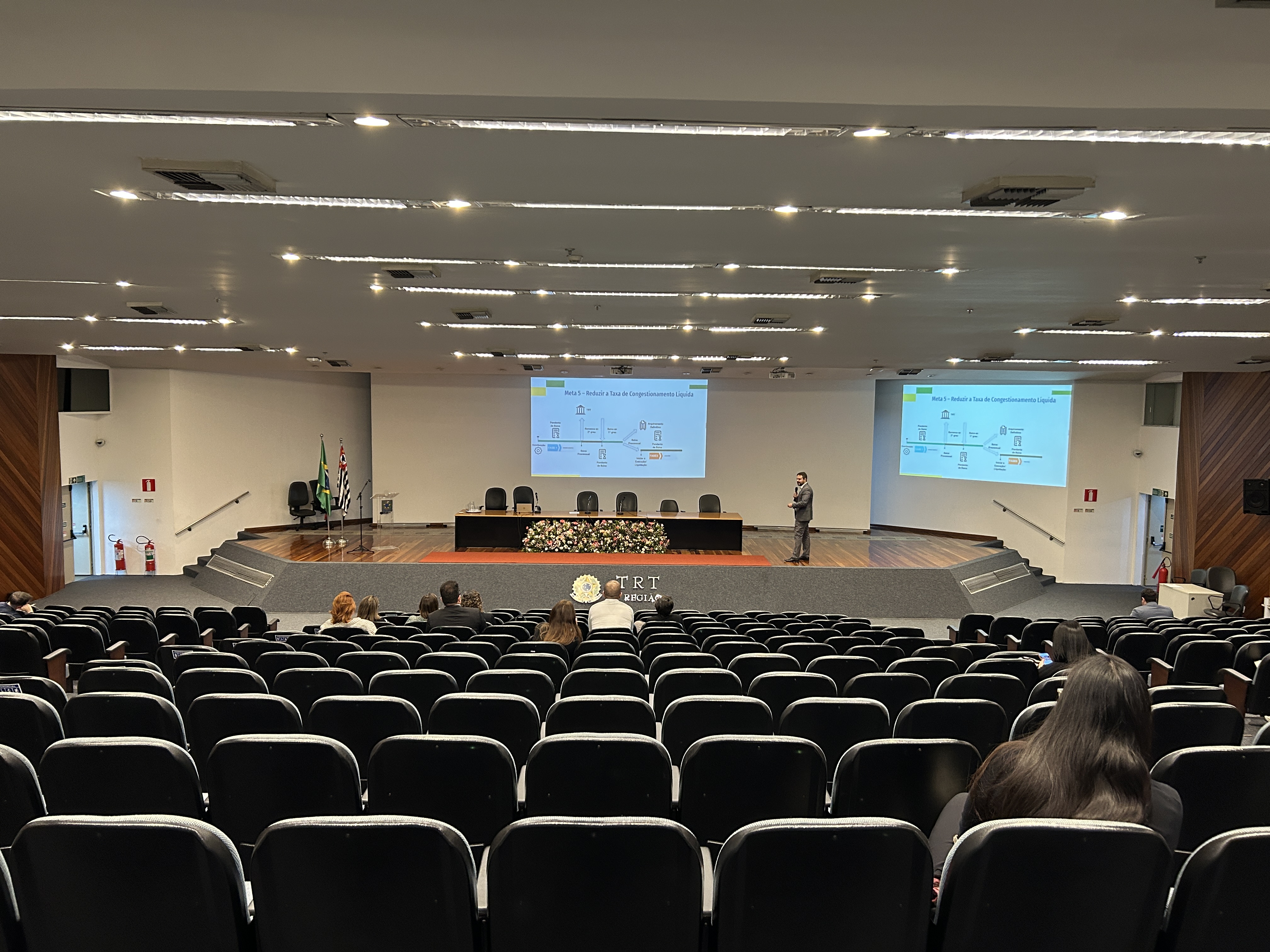 A imagem mostra um auditório com várias fileiras de cadeiras, onde ocorre uma apresentação. Um homem está em pé no palco, falando e apontando para dois telões com slides projetados. Há poucas pessoas assistindo, espalhadas pelas cadeiras. No palco, há uma mesa com arranjos florais e bandeiras do Brasil e do estado de São Paulo. O ambiente é formal e parece se tratar de uma palestra ou evento institucional no TRT (Tribunal Regional do Trabalho).